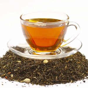 Een glazen kop gevuld met amberkleurige thee, geplaatst op een transparant schoteltje, rustend op een hoop losse zwarte thee gemengd met witte bloemblaadjes